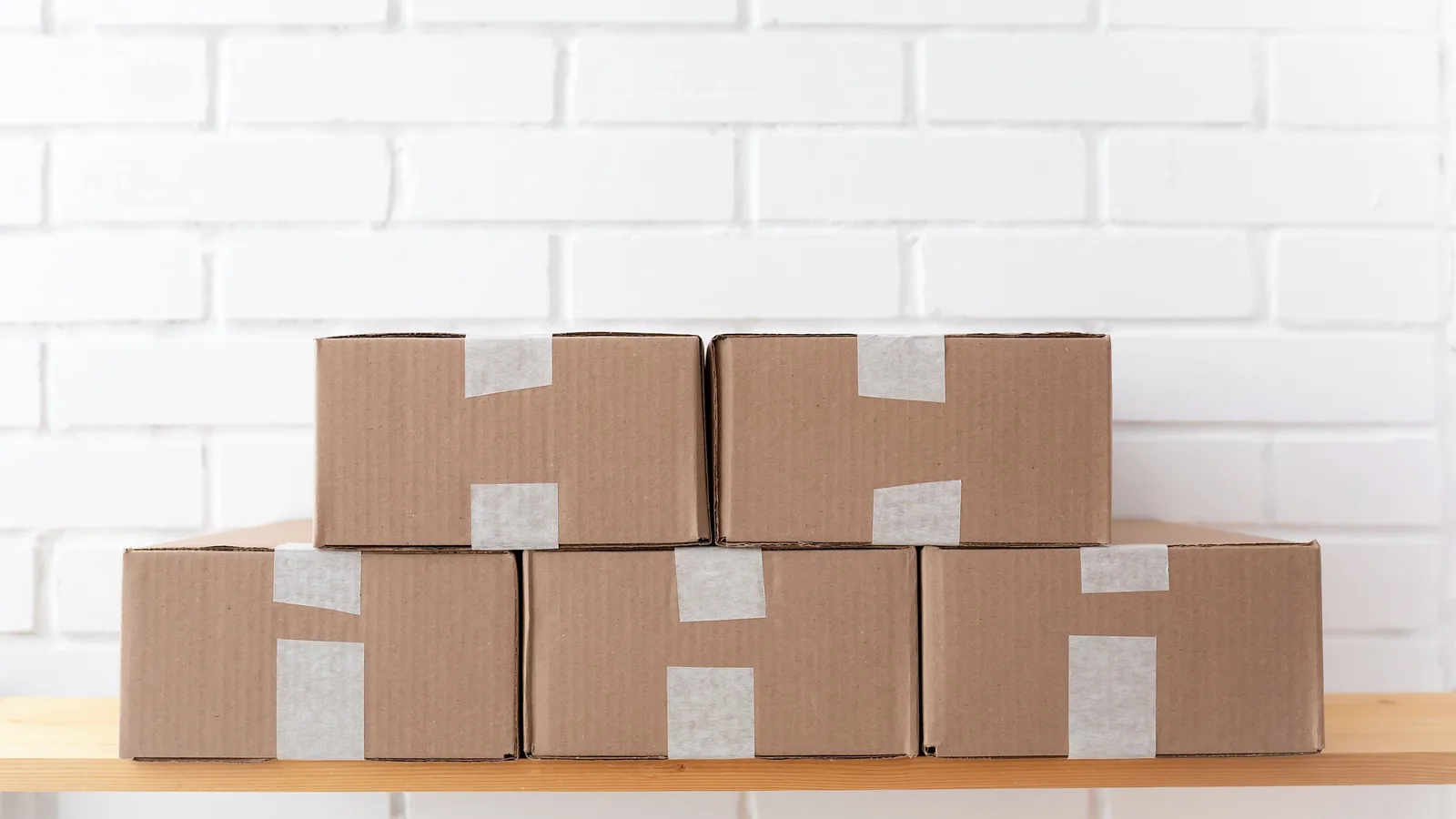 Taped cardboard storage boxes stacked on a wooden shelf against a white brick wall